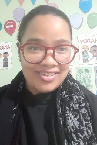 Smiling person wearing red-framed glasses and black jacket with patterned scarf against a colorful party-themed background