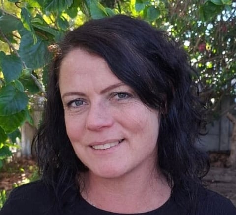 Woman with black wavy hair smiling at camera outdoors with green foliage in background