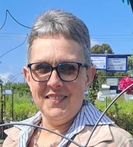 Middle-aged man with gray hair and glasses wearing a striped shirt outdoors with garden and greenhouse in background