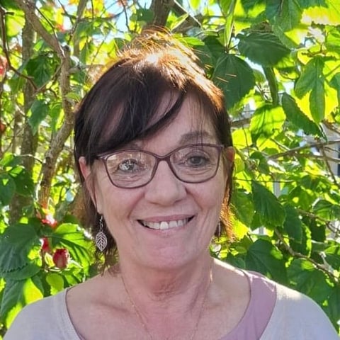 Portrait of a smiling woman with dark hair, glasses, and earrings in front of green foliage