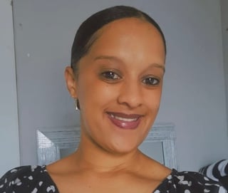 Portrait of a woman with short dark hair, wearing a black patterned top, smiling at the camera against a light blue wall background