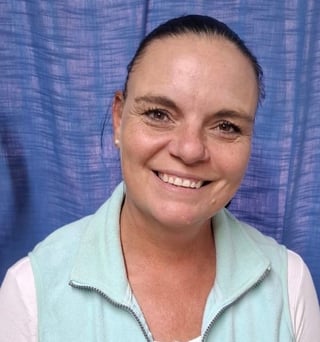 Smiling woman with dark hair pulled back wearing a light blue zip-up vest over a white shirt, posed against a blue textured background