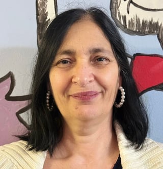 Middle-aged woman with long dark hair and pearl earrings smiling at camera, colorful artwork visible in background
