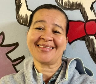 Middle-aged woman with short dark hair smiling at the camera wearing a light-colored jacket in front of colorful artwork on a wall